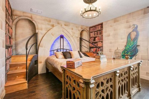 Cozy bedroom with a wooden bed, spiral staircase, and decorative bookshelves against a stone wall.