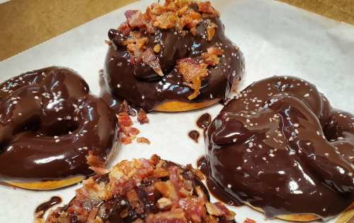 Three chocolate-glazed donuts topped with crispy bacon bits, served in a brown paper box.