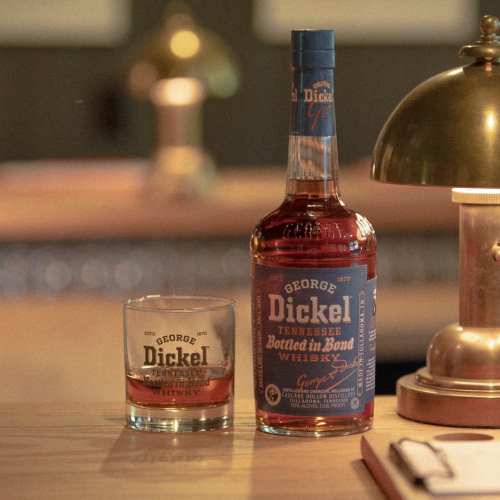 A bottle of George Dickel Tennessee whiskey next to a glass filled with whiskey, on a wooden table beside a lamp.