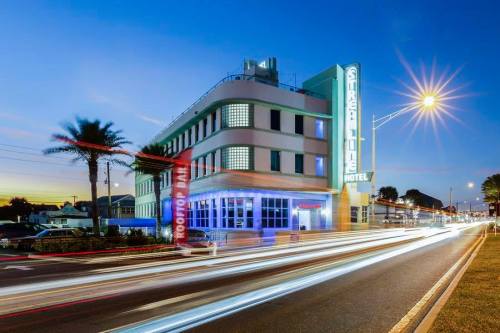 Art Deco hotel at dusk, featuring neon lights and palm trees, with light trails from passing cars on the road.