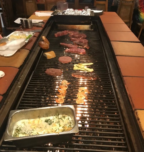Grill with various meats, including steaks and shrimp, cooking over an open flame at a restaurant.