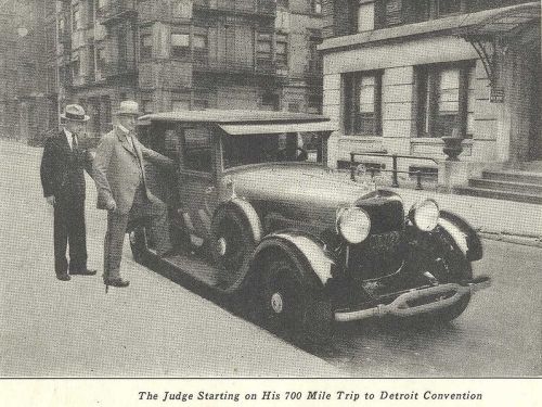 A vintage car with two men, one standing beside it, preparing for a 700-mile trip to a convention in Detroit.