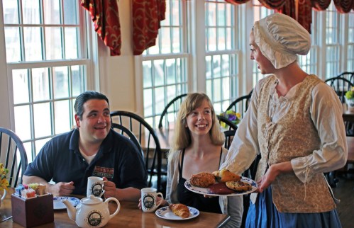 A server in period costume presents pastries to two smiling diners at a cozy restaurant with large windows.