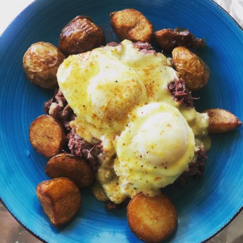 A blue bowl filled with poached eggs on corned beef, topped with hollandaise sauce, and surrounded by roasted potatoes.