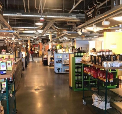A spacious market interior with shelves of products, including wine and snacks, and shoppers in the background.