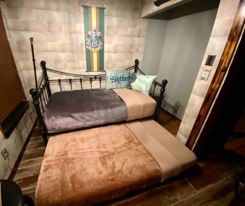 A cozy bedroom with a black metal bed, two different colored blankets, and a Slytherin-themed pillow against a stone wall.