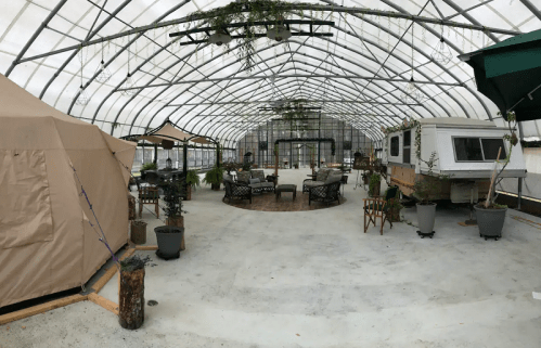 A spacious greenhouse with seating areas, plants, and a vintage trailer, creating a cozy, inviting atmosphere.