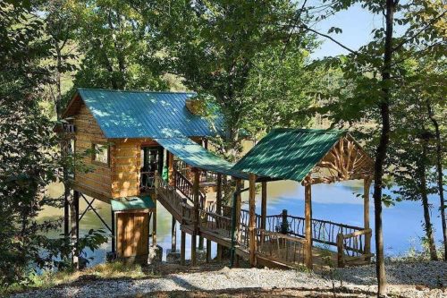 A wooden treehouse with a green metal roof, surrounded by trees and overlooking a calm river.