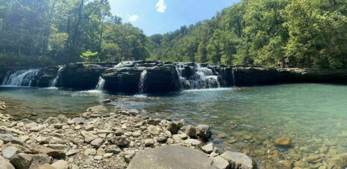 A serene waterfall cascades over rocks into a clear, turquoise pool, surrounded by lush green trees and a blue sky.