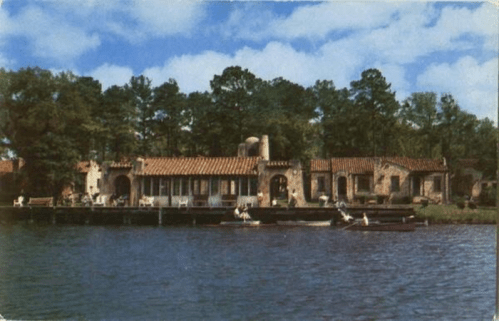 A scenic view of a rustic building by a lake, with kayakers and lush trees under a blue sky.
