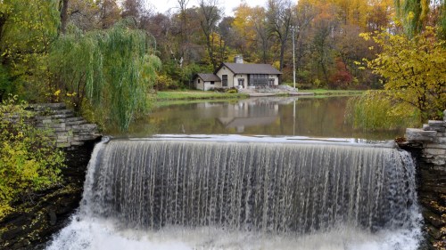 A serene scene featuring a waterfall in the foreground, with a house and colorful autumn trees in the background.