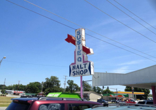 Sign for "Susie Q Malt Shop" with a bright blue sky and parked cars in the foreground.