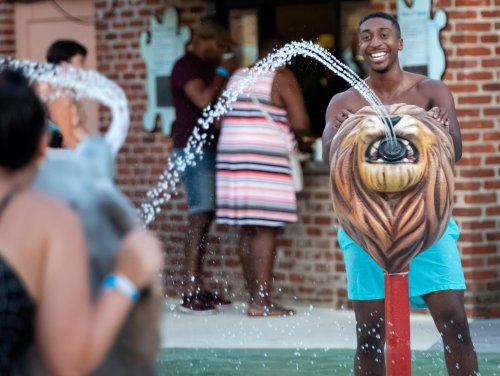 A man smiles while playfully interacting with a lion-shaped water fountain, with others in the background.