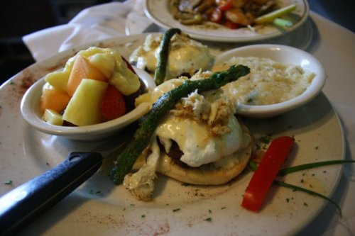 A plate featuring eggs Benedict with crab, asparagus, grits, and a fruit medley on the side.