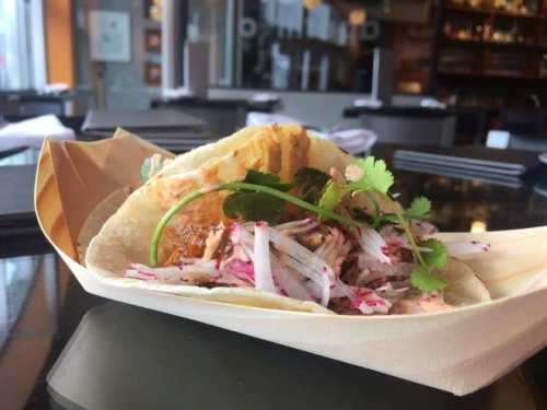 A taco filled with shredded meat, topped with radishes and cilantro, served in a paper holder on a table.