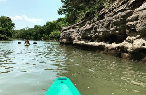 A person kayaking on a calm river, surrounded by lush greenery and rocky cliffs under a clear blue sky.