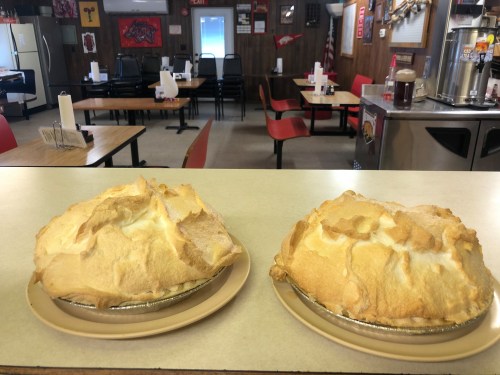Two golden meringue pies on plates, set on a table in a cozy diner with empty chairs and decor in the background.