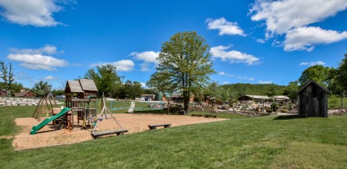 A sunny playground with swings and a slide, surrounded by green grass and trees, under a blue sky with fluffy clouds.