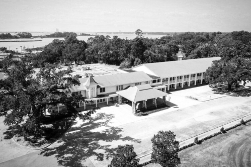 Aerial view of a large, two-story building surrounded by trees and a body of water in the background, in black and white.