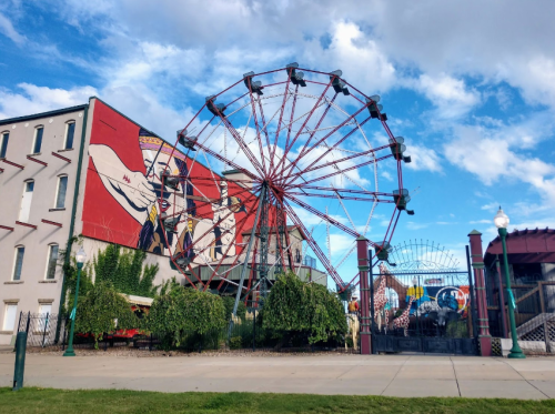 A colorful mural on a building beside a large, red Ferris wheel under a blue sky with scattered clouds.