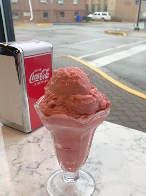 A glass dish filled with pink strawberry ice cream sits on a marble table, with a Coca-Cola dispenser in the background.