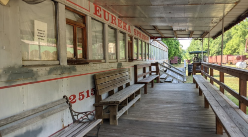 A wooden porch with benches in front of a vintage train car, labeled "Eureka Springs," surrounded by greenery.
