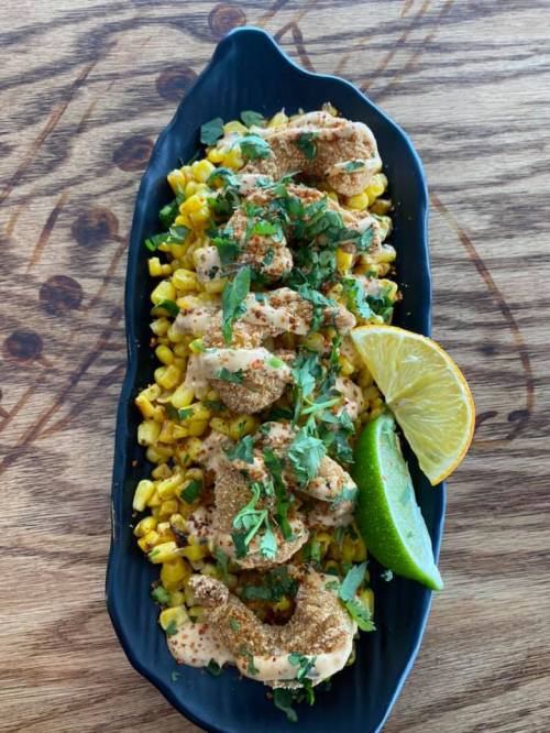 A black platter with shrimp on a bed of corn, garnished with herbs and lime wedges.