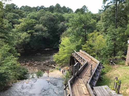 A scenic view of a river surrounded by trees, with a wooden structure and people near the water's edge.
