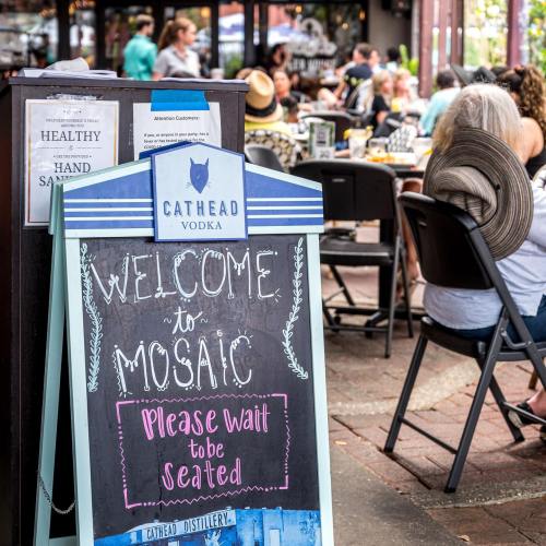 A sign reading "Welcome to Mosaic" at a bustling outdoor dining area with people seated at tables.