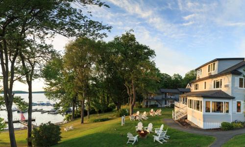 A scenic lakeside view featuring a white house, green lawn, and boats docked in the water under a clear sky.