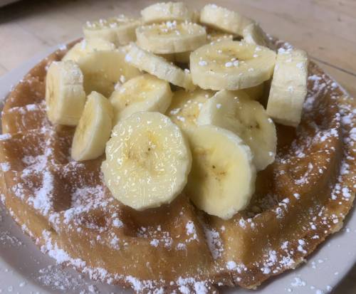 A golden waffle topped with sliced bananas and dusted with powdered sugar on a white plate.