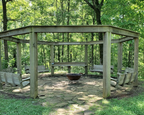A circular wooden gazebo surrounded by benches, with a fire pit in the center, set in a lush green forest.