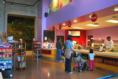A colorful cafeteria with a food counter, customers ordering, and a child in a stroller. Snacks are displayed on the side.