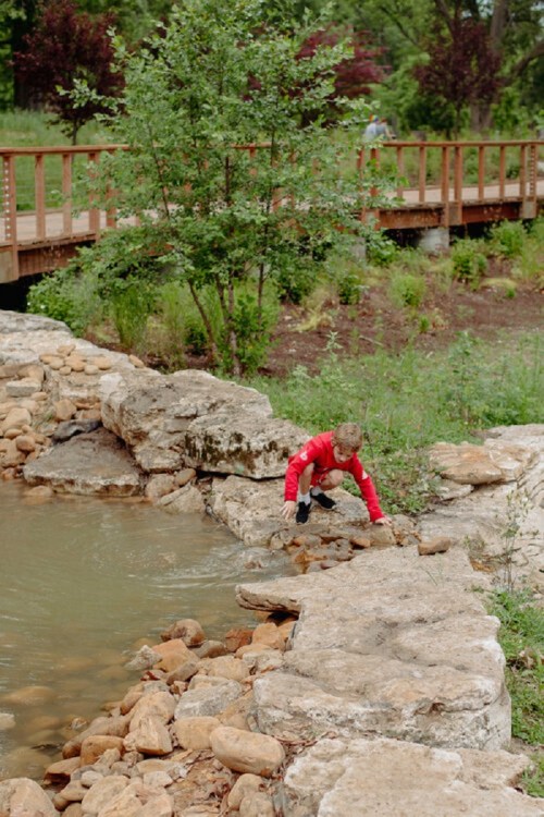 A child in a red shirt climbs over rocks by a small pond, with a wooden bridge and greenery in the background.