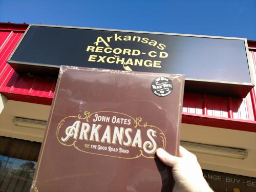 A person holds a vinyl record titled "Arkansas" in front of the Arkansas Record-CD Exchange store sign.