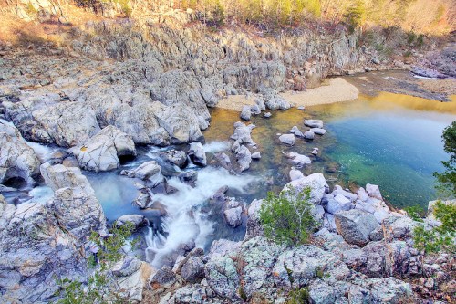 A serene river flows over rocky terrain, surrounded by trees and a calm pool of water reflecting the landscape.