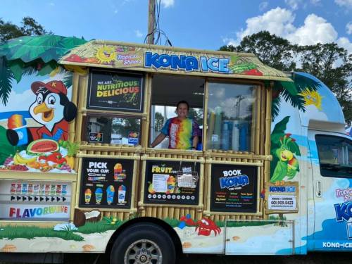 A colorful Kona Ice truck with a smiling employee in a tie-dye shirt, offering shaved ice and tropical treats.