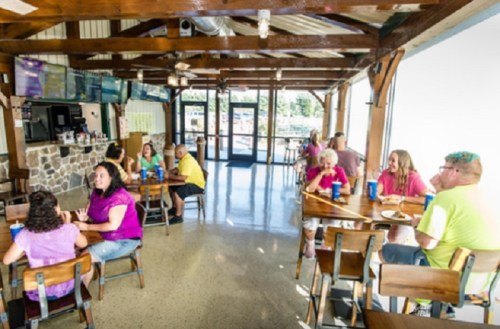 A lively restaurant interior with patrons enjoying food and drinks at wooden tables, featuring a stone bar and large windows.