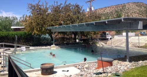 A serene outdoor pool area with people swimming, surrounded by trees and mountains under a shaded structure.