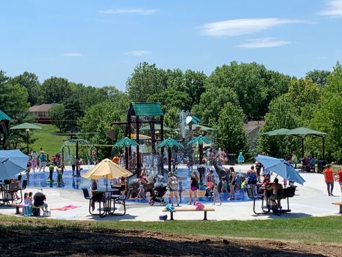 A vibrant splash pad filled with children playing, surrounded by trees and umbrellas on a sunny day.