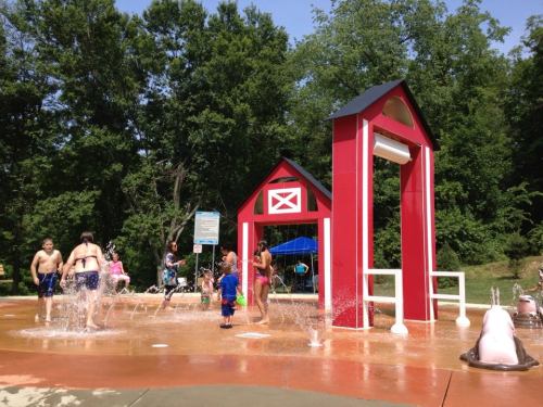 Children play in a splash pad featuring a red barn structure, surrounded by trees on a sunny day.