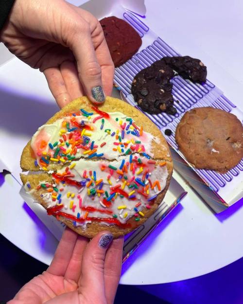 A hand holds a large frosted cookie topped with colorful sprinkles, surrounded by other assorted cookies on a table.