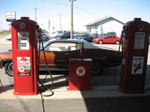 Vintage gas station with red pumps and a classic brown car parked nearby.