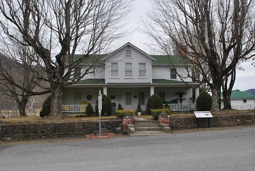A two-story white house with a green roof, surrounded by bare trees and a stone wall, set in a rural landscape.