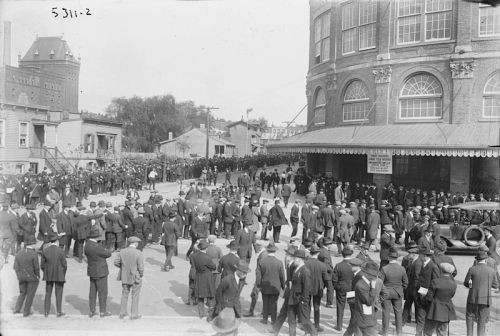A large crowd of people in formal attire gathers outside a historic building, with some onlookers and vehicles nearby.