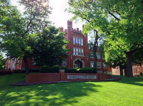 A red brick building with a tower, surrounded by green trees and grass, featuring a sign that reads "Marshall University."