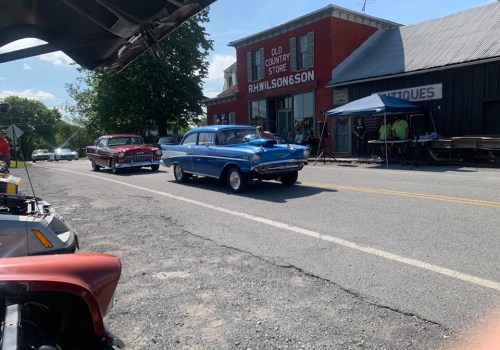Two classic cars drive past an antique store on a sunny day, with people gathered under a tent nearby.