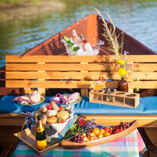 A cozy picnic setup on a boat, featuring fruits, pastries, drinks, and flowers, with a scenic water view.