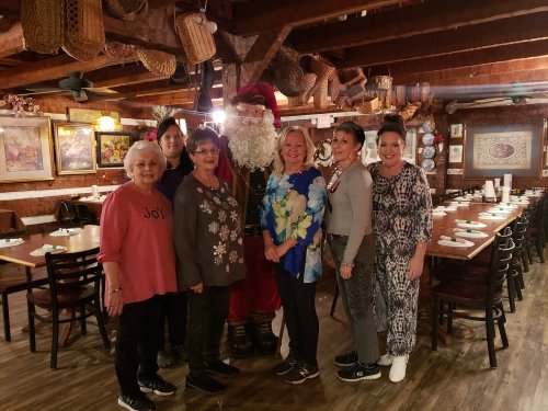 A group of six women poses with Santa in a cozy restaurant decorated with rustic elements.