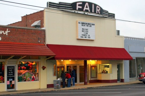 A vintage theater with a marquee sign reading "FAIR" and a crowd outside, promoting a music show for charity.